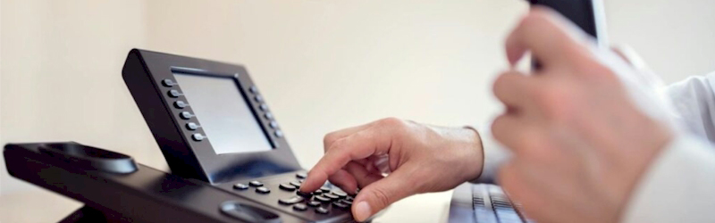 Hand pressing buttons on a modern VoIP desk phone with a digital display.