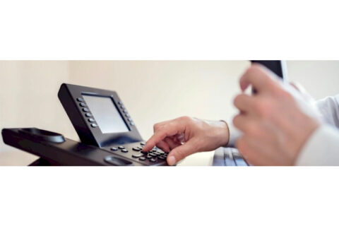 Hand pressing buttons on a modern VoIP desk phone with a digital display.